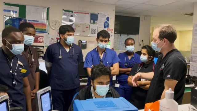 Workers at a hospital are wearing uniforms and face masks while standing in a circle talking to one another.