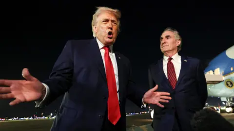 Reuters US President Donald Trump on the airport tarmac with Air Force One behind him. The president has his hands and mouth open as he speaks to a boom mic. Secretary of the Interior Doug Burgum stands to the president's left and watches him speak. 