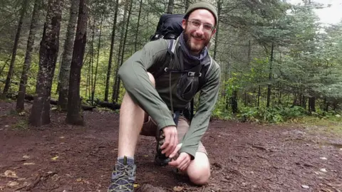 Family handout Alex is pictured in woodland wearing shorts and walking boots while kneeling on the ground 