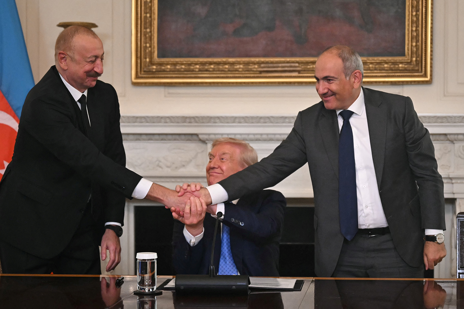 Trump, Azerbaijani President Ilham Aliyev, and Armenian Prime Minister Nikol Pashinyan shake hands simultaneously in a room with a white marble fireplace and a gold-framed painting. A document rests on a desk in front of them.