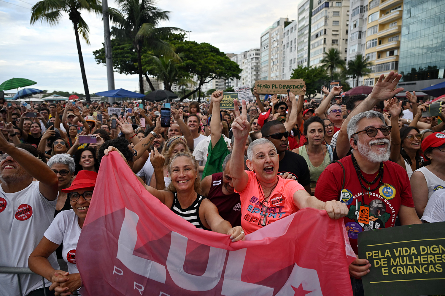 A large crowd of people gathers at a beachfront, with participants holding a pink banner reading "Lula" and raising their hands.