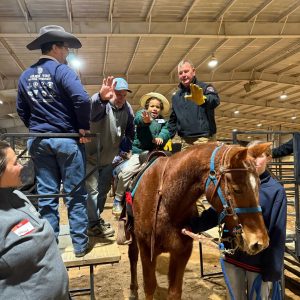 A child sits on a horse while nearby adults wave inside an indoor riding arena.