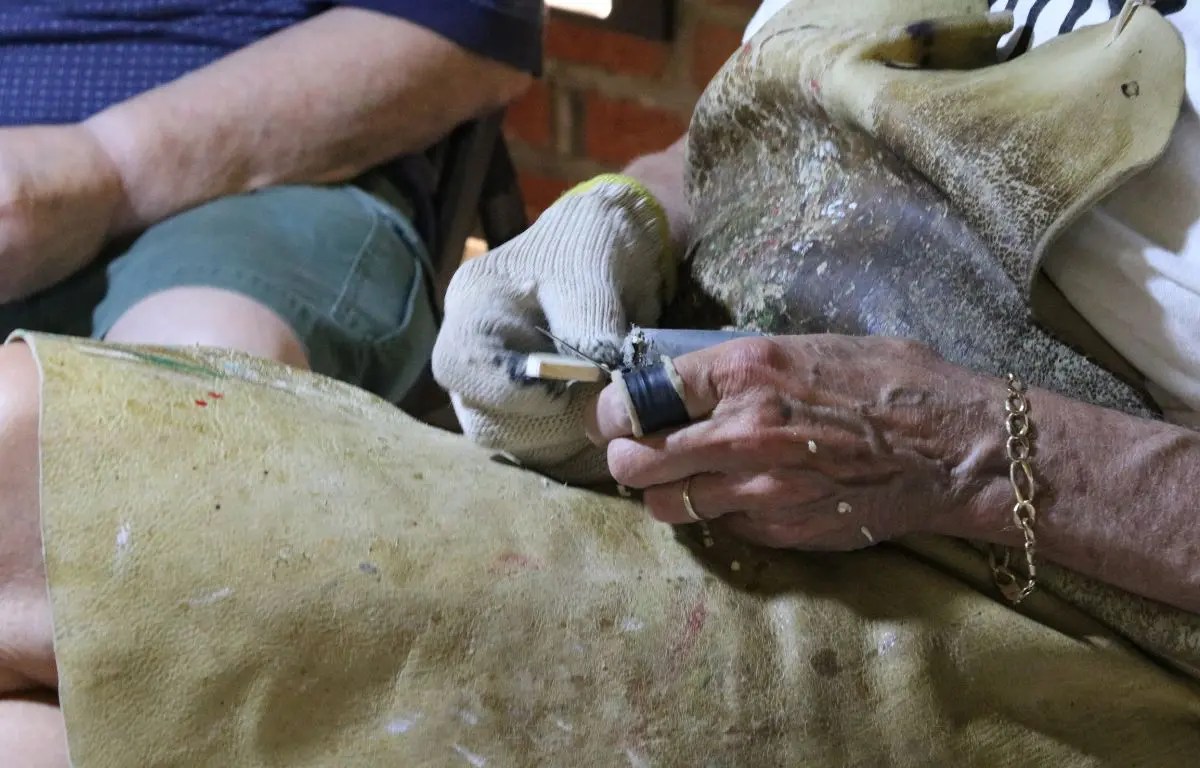 A man holds a wood block with a gloved hand, carving it with a tool in an uncovered hand while wearing an apron.