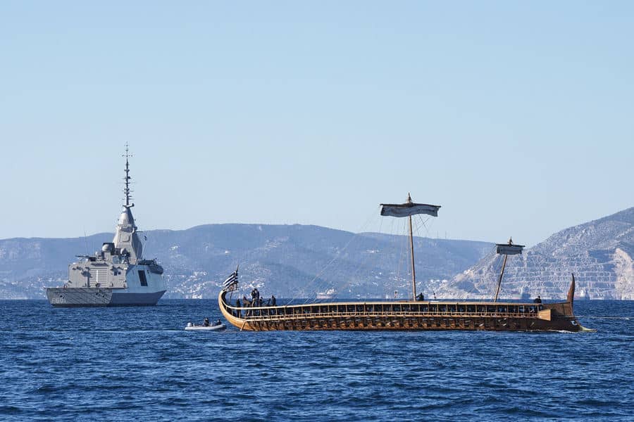 A modern grey naval frigate, the Kimon (F601), is positioned in the background of blue coastal waters, while a wooden Ancient Greek trireme, the Olympias, sails in the foreground, showcasing two eras of Greek maritime history. What is the relationship between and thalassocracy?