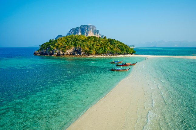 A high angle view of Krabi beach, in Thailand, with three wooden boats on the water and a tropical island in the background.