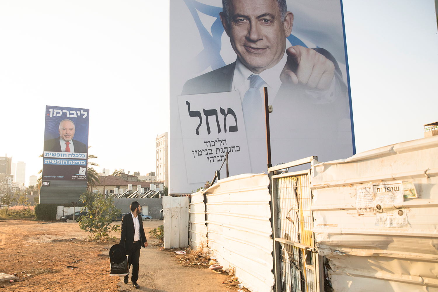 An Orthodox Jewish man in traditional black attire walks past large campaign billboards in Israel. The largest billboard depicts Israeli Prime Minister Benjamin Netanyahu pointing.