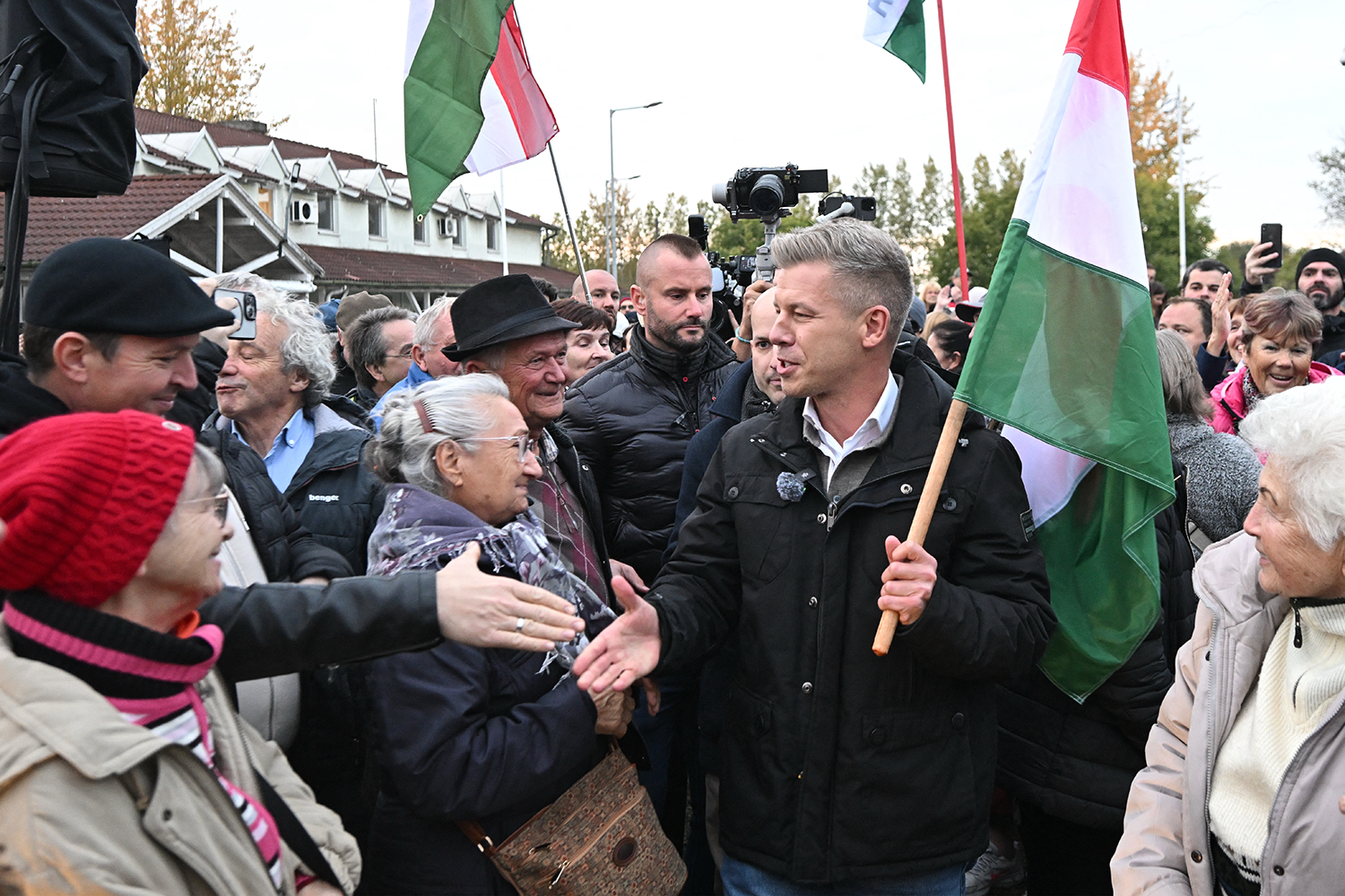 Peter Magyar, wearing a dark jacket and carrying a Hungarian flag, interacts warmly with a crowd of people while surrounded by media cameras.