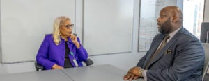 A woman in a purple blazer and glasses listens thoughtfully during a meeting across a conference table from a man in a dark suit, in a bright office with whiteboards in the background.