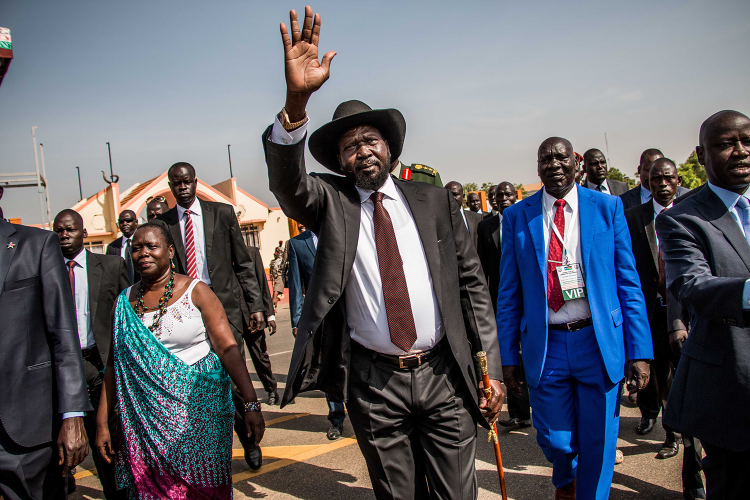 A political leader wearing a black fedora hat and dark suit waves to a crowd while walking with an entourage of officials and security personnel.
