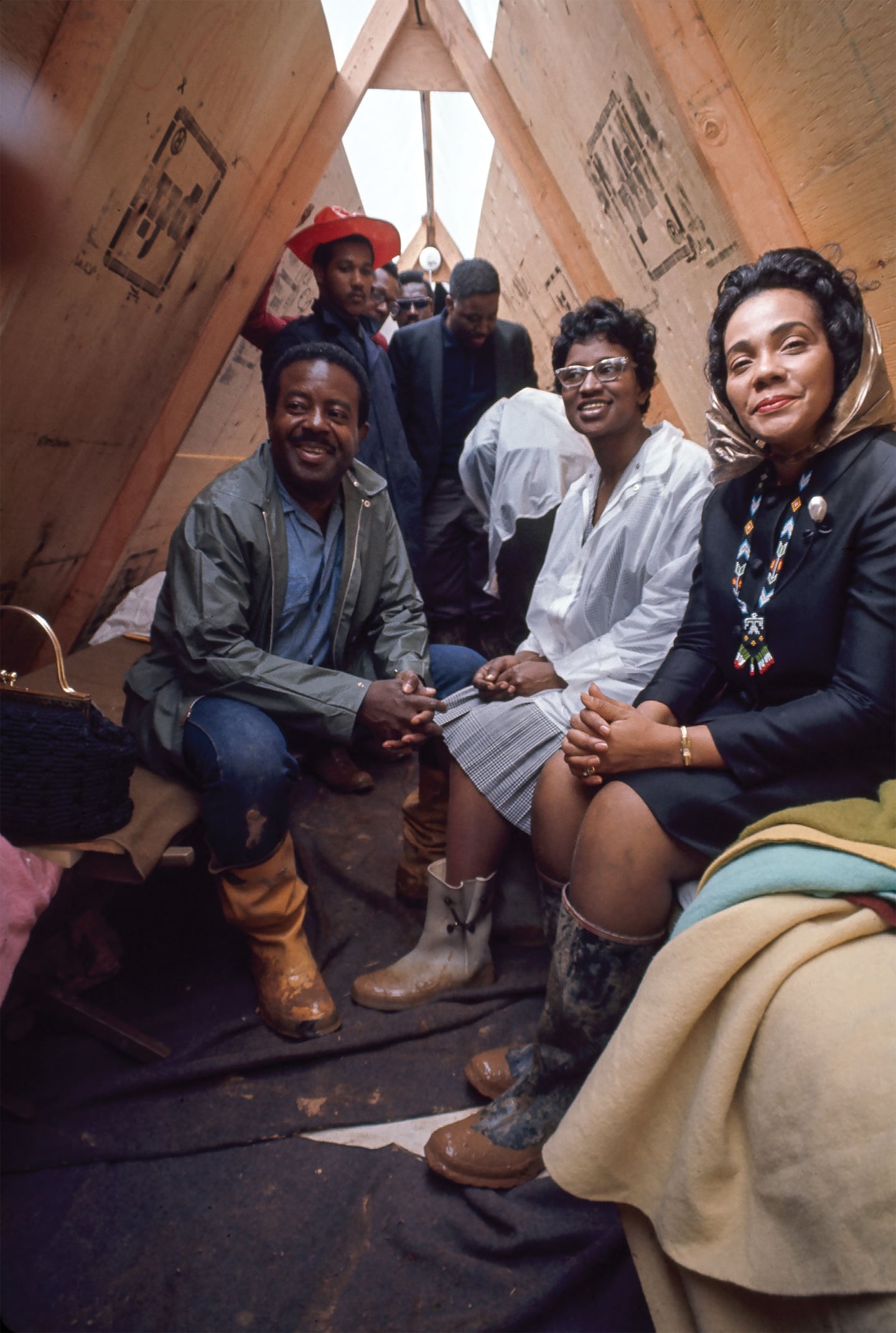 A group of people sitting inside a wooden shelter, wearing rain boots and coats, with a woman in a headscarf smiling at the camera.