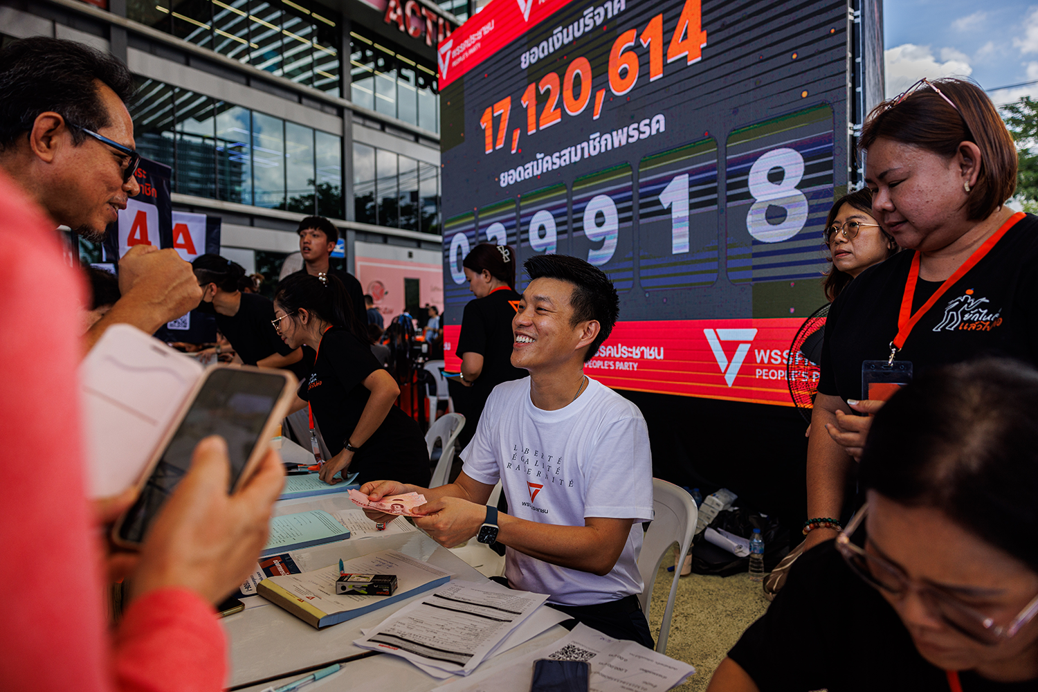A smiling man in a white t-shirt sits at a table accepting donations. A large electronic display board shows the numbers "17,120,614" and "609918."