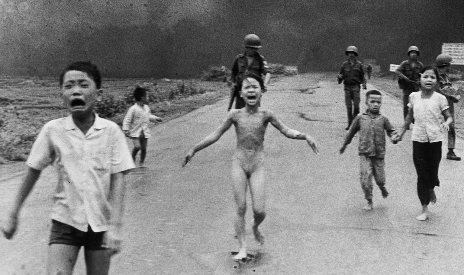 A group of children, some crying and barefoot, run down a road toward the camera with smoke and armed soldiers in the background. The central child appears distressed and unclothed.