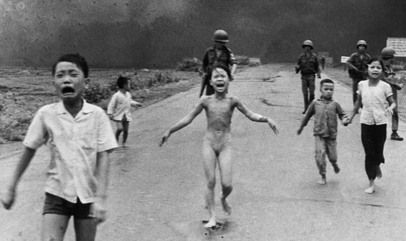 A group of children, some crying and barefoot, run down a road toward the camera with smoke and armed soldiers in the background. The central child appears distressed and unclothed.
