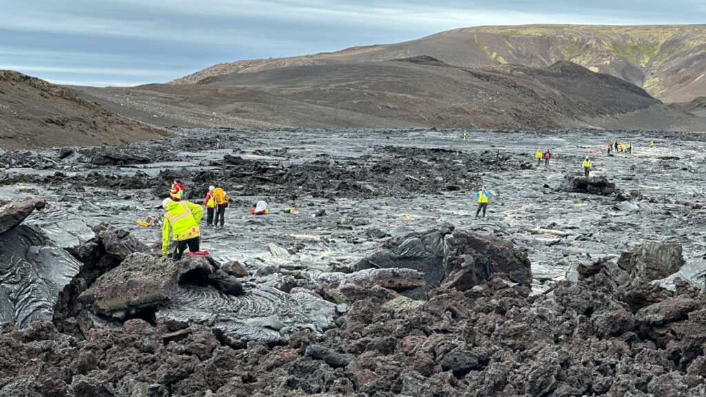 Discovering Venus on Iceland – Eos Scientists wearing bright yellow safety vests stand in various places amid an expanse of dark volcanic rock with barren hills in the background.