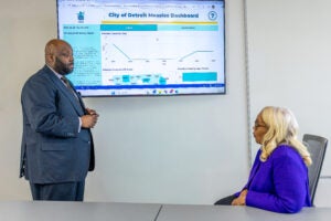A large man in a suit stands before a presentation screen displaying the City of Detroit measles dashboard facing a woman in a business suit seated at a conference room table.
