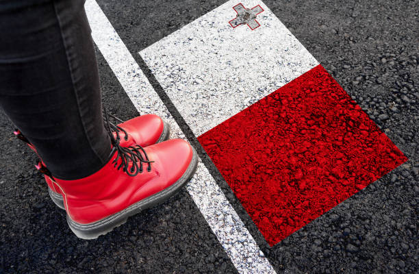Person wearing red boots stands next to a Malta flag painted on asphalt near a road marking.