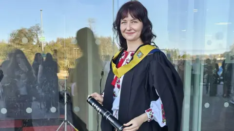 Svitlana Savinova Svitlana in an academic robe and sash in front of a modern building. She is holding a certificate in a black cardboard tube. She is smiling and has dark hair.