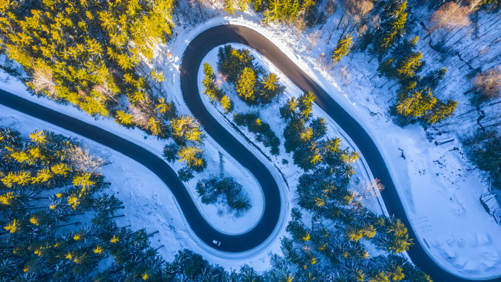 Asphalt road serpentine in snowy wintertime. Cold winter and sunny day above forest road with illuminated trees by rising sun. Aerial view from drone.