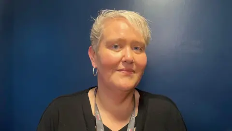 Adele McBurney. She has short blonde hair and blue eyes, wearing a black top, silver hoop earrings and a grey lanyard. A dark blue wall is behind her.