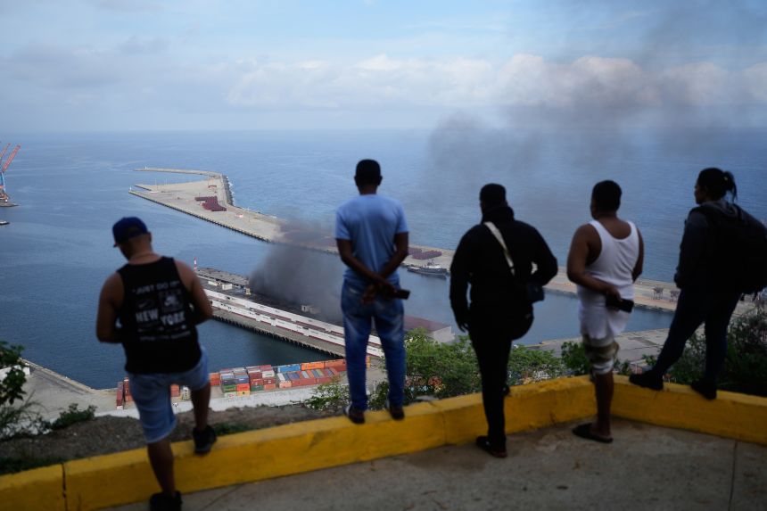 Men watch smoke rising from a dock after explosions were heard at La Guaira port, Venezuela, Saturday, on January 3, 2026.
