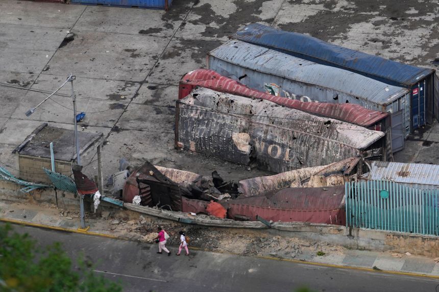 Pedestrians walk past destroyed containers lay at La Guaira port after explosions were heard in Venezuela, Saturday, Jan. 3, 2026.