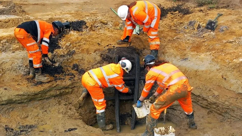 Four archaeologists in hi-vis orange suits and black boots bending over a sandy soiled stepped site. In the middle is a blackened ladder with three rungs emerging from a watery pit.