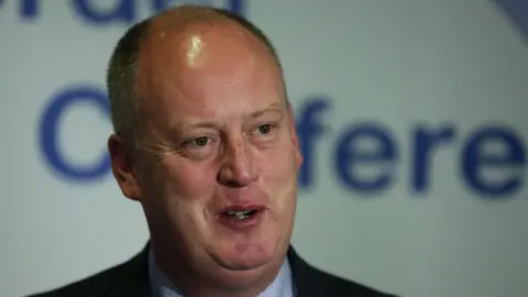 PA Media Sir George Hamilton at a conference. He is wearing a navy suit and blue shirt. He has short grey hair. 