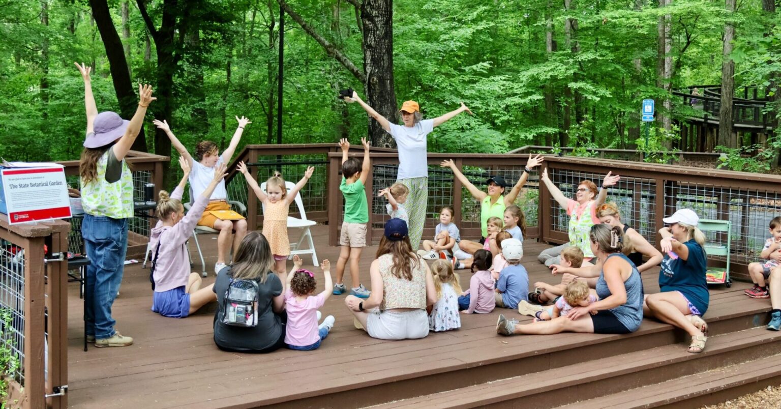 A group of adults and children put their arms in the air while sitting on a platform in the woods.