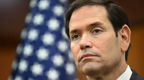 Getty Images An image of the US Secretary of State, Marco Rubio, speaking into a microphone while wearing a navy suit with a red tie.
