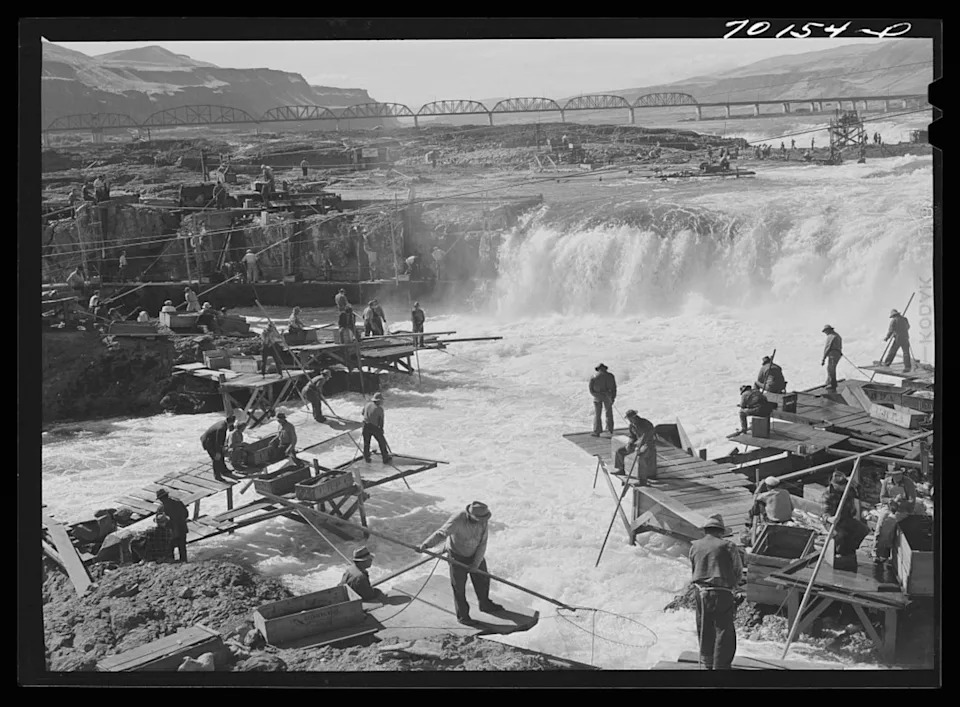 People stand on rocks and wooden platforms near a rocky waterfall, fishing.