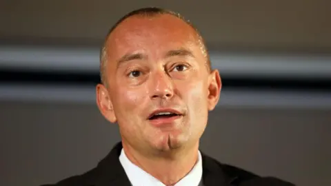 Getty Images Bulgarian politician Nickolay Mladenov speaking into a microphone while dressed in black suit and pale blue tie. 
