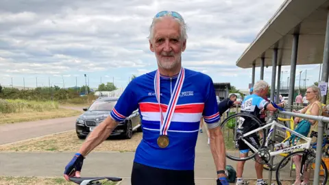 Brian Cookson Brian Cookson stands next to his bike after cycling in Mallorca in 2025. He is wearing a blue cycling top and shorts, and has a medal around his neck. 