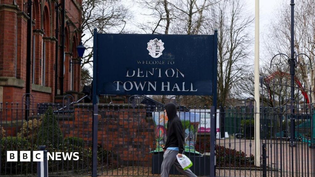 A big blue sign says Welcome to Denton Town Hall outside a redbrick historic building. A man in a hooded top with carrier bag walks past it.