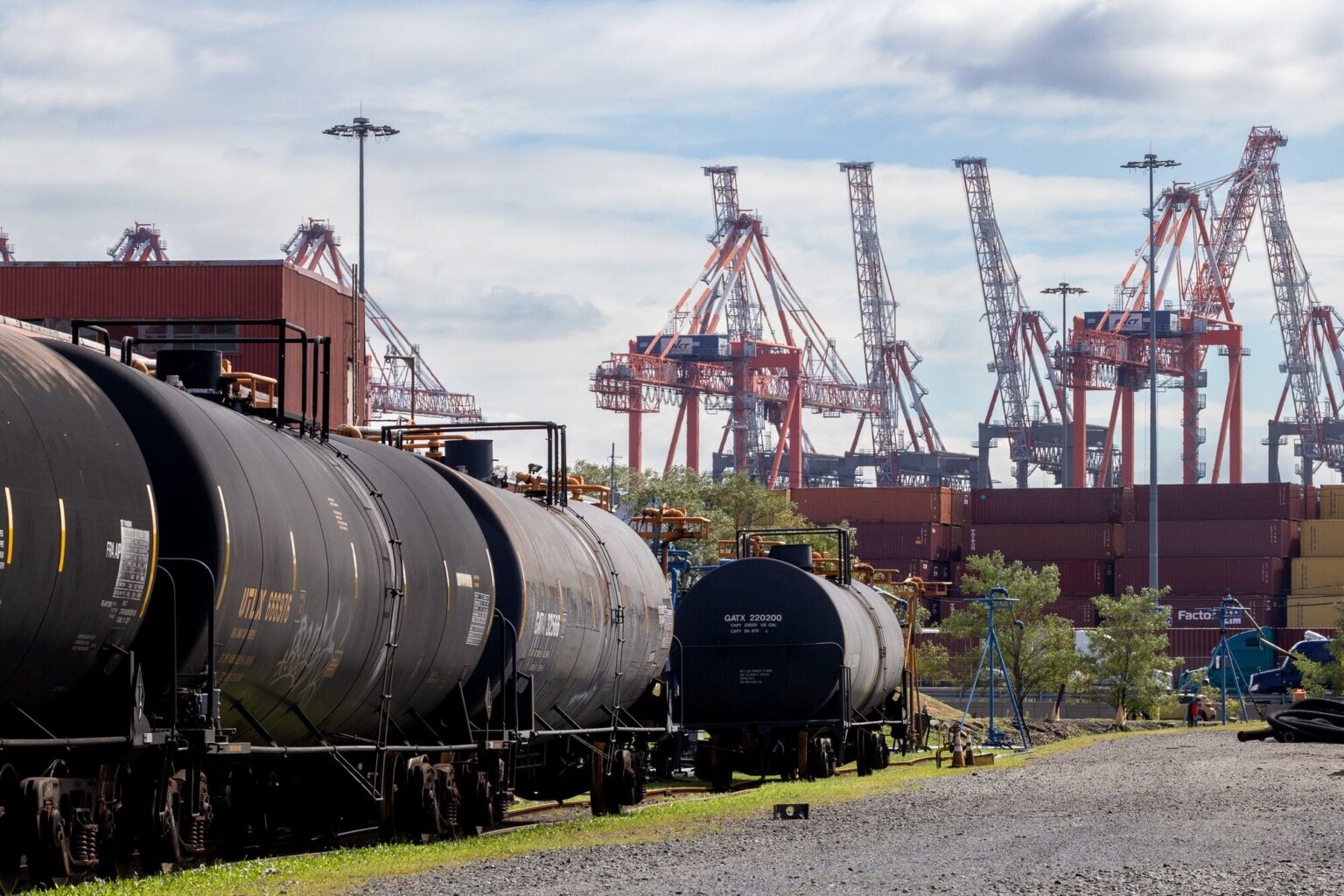 Black, cylindrical train cars on tracks next to stacks of containers and large cranes in the distance.