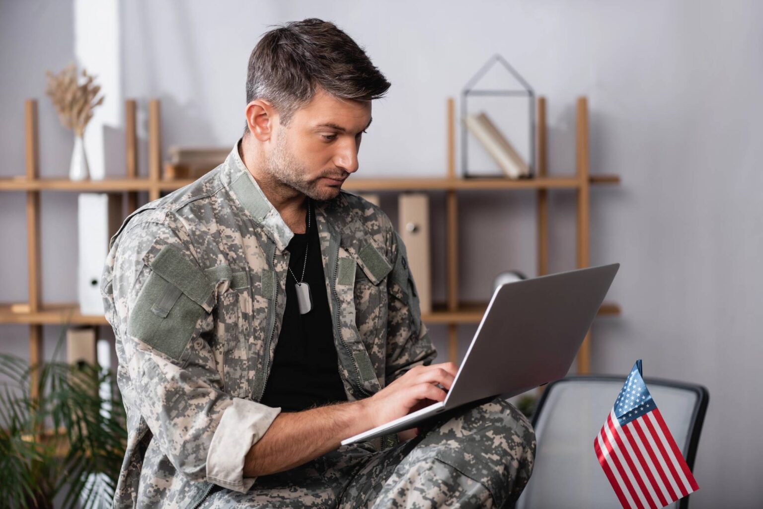 Can I Move to Portugal on VA Disability Compensation? A man in a camouflage uniform and dog tags sits indoors using a laptop, with an American flag nearby; shelves are in the background.