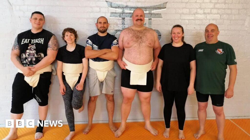 A sumo referee and Jones on a raised ring during a match. Jones is positioned in a low starting stance, with both hands touching the ground. She is wearing a black athletic outfit along with a traditional mawashi tied at the back. The referee, dressed entirely in white, is leaning forward as if preparing to signal the start of the bout. In the background, an audience is seated in a large indoor venue, watching the action unfold.