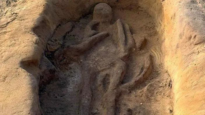 Shadow skeletons of an adult and child emerging from sandy soil in a pit. The head of the adult is at the top and its arms, torso and legs can be seen. The bod of the  child is tucked in at its right side, with its left arm folded over its body.