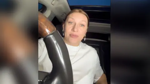 Telegram Polina Azarnykh in a white T-shirt holding a large steering wheel, looking directly at the camera. Her fair hair is pulled back from her face.