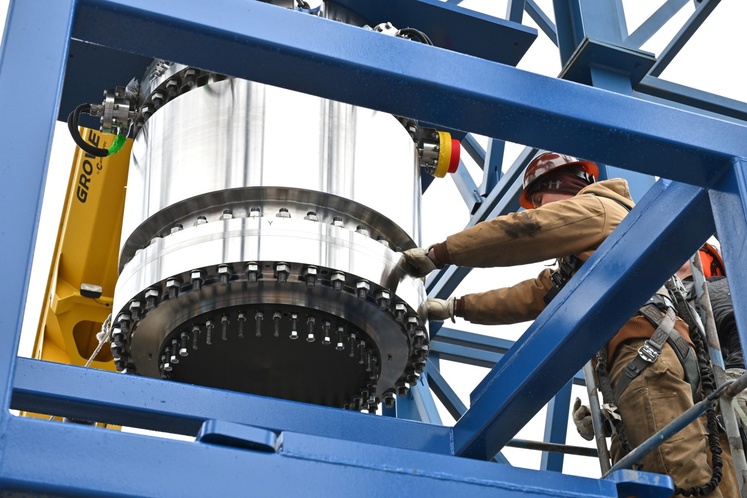 Workers in safety gear guide a large white cylindrical rocket motor or engine component with a black mounting flange as it's positioned within a blue steel test stand structure. Yellow crane equipment is visible on the left side