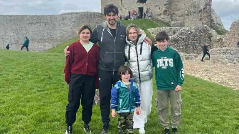 Alex Sivko The family are standing in a row in leisure wear in front of the ruins of a castle. They are smiling for the camera and dad Alex has his arms around his wife and two oldest children while the youngest stands in front of them. 