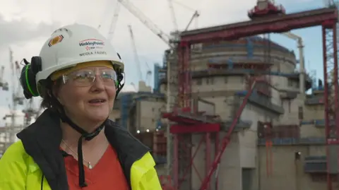 BBC Nicola Fauvel is standing in front of the Hinkley plant that is under construction. She is wearing a red jumper, hi-vis jacket, safety goggles and a white hard hat with her name on. She also has earphones resting on the helmet. There are numerous cranes in the background.