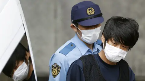 Reuters Tetsuya Yamagami, suspected of killing former Japanese Prime Minister Shinzo Abe, is escorted by a police officer as he is taken to prosecutors, at a police station in Nara in 2022.