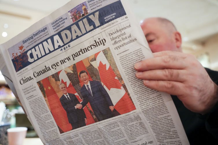 A man reading a newspaper with a picture of Mark Carney and Xi Jinping shaking hands on the front