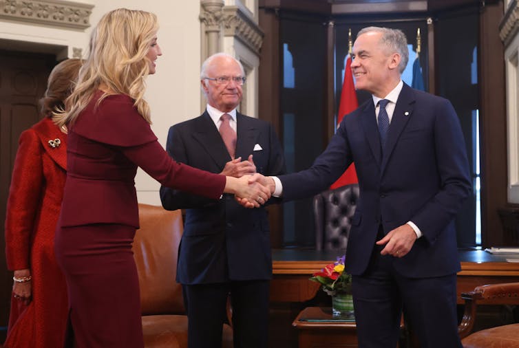 A tall blonde woman in a burgundy dress shakes hands with a thin smiling man with short grey hair.