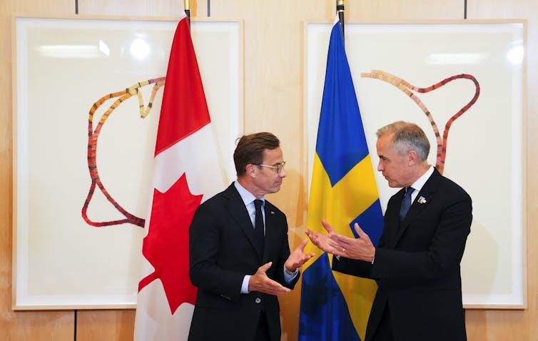 Two men in suits stand talking in front of the Canadian and Swedish flags.