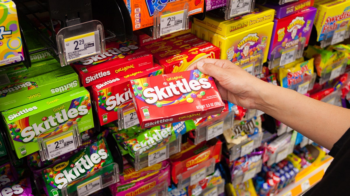 Person's hand seen buying skittles from packed and colorful candy display in store.