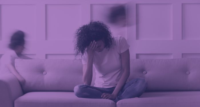 a brown-skinned woman sitting cross-legged on a couch with her head in her hands while two children are seen blurred as through running in the background