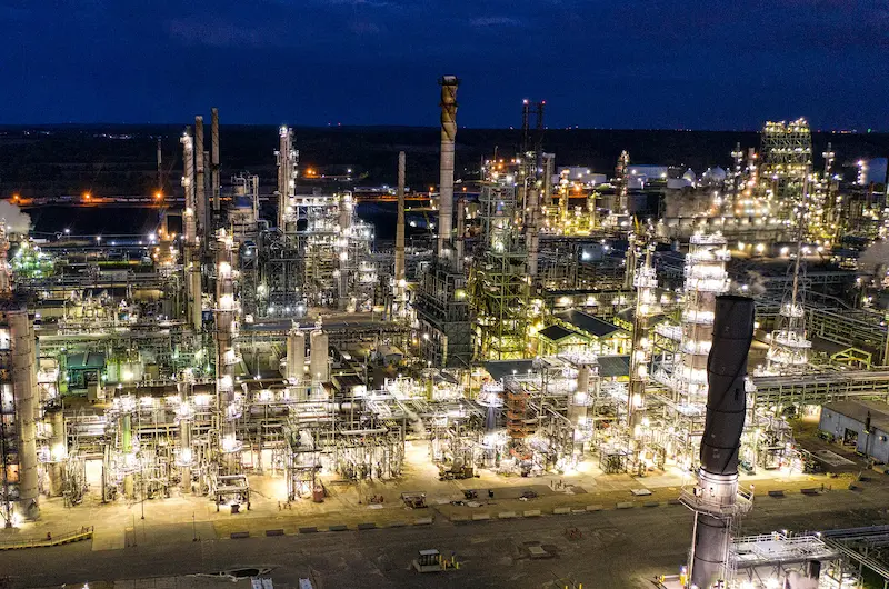 An aerial view of a vast industrial refinery, brightly lit at night. Thousands of yellow lights illuminate the intricate network of towering columns, pipes, and buildings under a dark blue sky, with visible steam plumes.