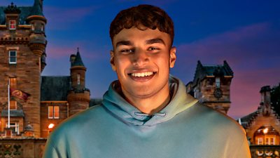 A young man in a light blue hoodie smiles to camera, set against the backdrop of a castle by night