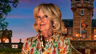 A woman in a patterned blouse smiles to camera, standing in front of a castle by night